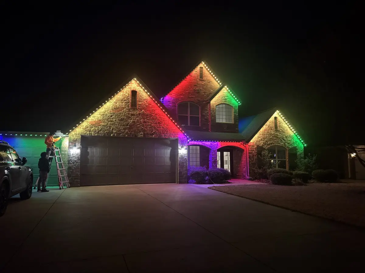 Peak installer on a ladder fitting lights to the roofline of a home lit in red, green and blue