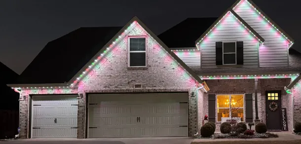 Brick ranch front at night in pink and green holiday lighting with a wreath on the door