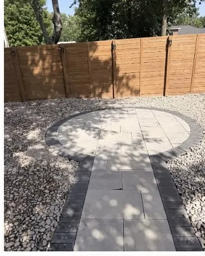 Circular paver patio set into river rock in the shade of a tree beside a wood fence