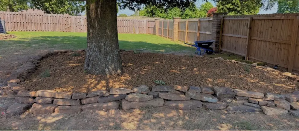 Stacked natural stone retaining wall holding a mulch bed around a mature tree in a fenced backyard