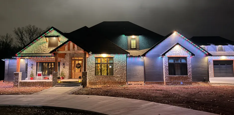 Stone and board front on a large home at night with red, green and white holiday lighting along every gable